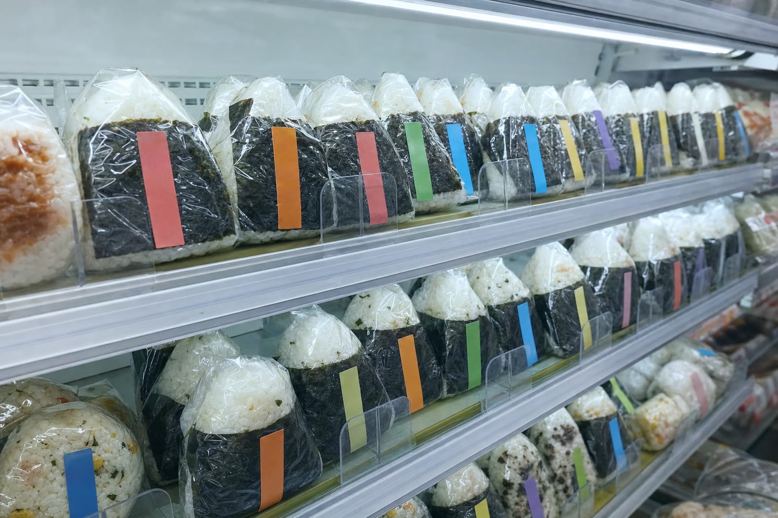A convenience store onigiri rice ball shelf in Japan, triangular rice balls wrapped in plastic film with colored flavor labels